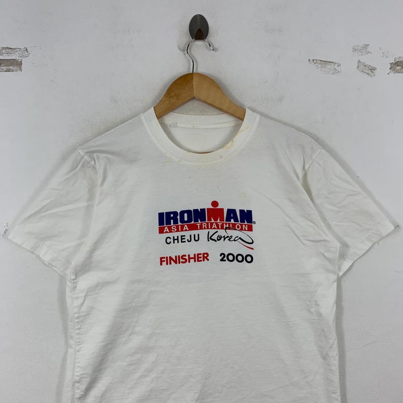 K&ouml;nnte beinhalten: Ein wei&szlig;es T-Shirt mit kurzen &Auml;rmeln und einem Aufdruck auf der Brust mit der Aufschrift IRONMAN ASIA TRIATHLON CHEJU Korea FINISHER 2000. Der Text ist in Blau, Rot und Schwarz gedruckt, wobei das Ironman-Logo &uuml;ber den Veranstaltungsdetails steht.