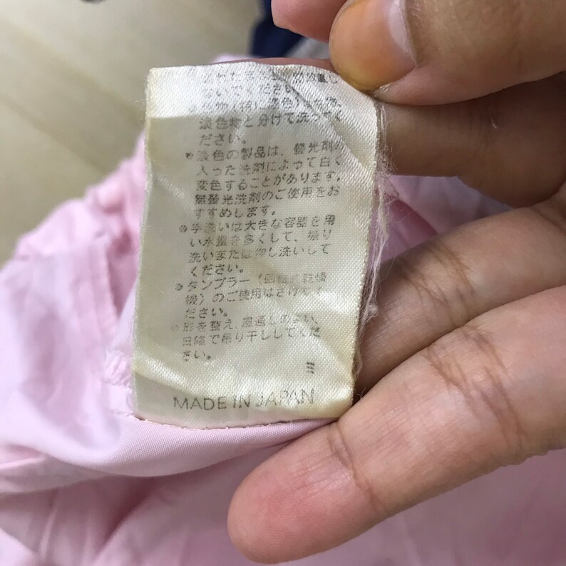 May include: A close-up of a beige tag with Japanese text and the words "MADE IN JAPAN" in black. The tag is attached to a pink fabric. The tag has a slightly wrinkled appearance.