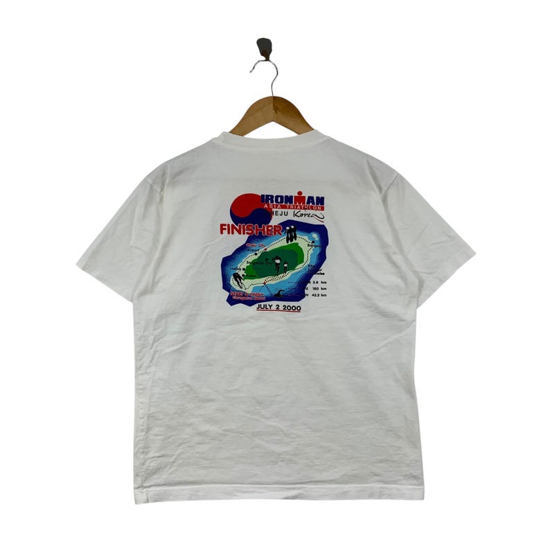 K&ouml;nnte beinhalten: Ein wei&szlig;es Kurzarm-T-Shirt mit einem Aufdruck auf der R&uuml;ckseite f&uuml;r den Ironman Asia Triathlon Jeju Korea. Das Design zeigt eine Karte der Insel, den Schriftzug FINISHER, das Datum JULY 2 2000 sowie Angaben zur Renndistanz in Kilometern.