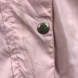 May include: Close-up of a pale pink jacket with a silver button. The button has the text "Sportswear" and the letters "AG" in a heart shape. The jacket has a vertical seam and some stains.