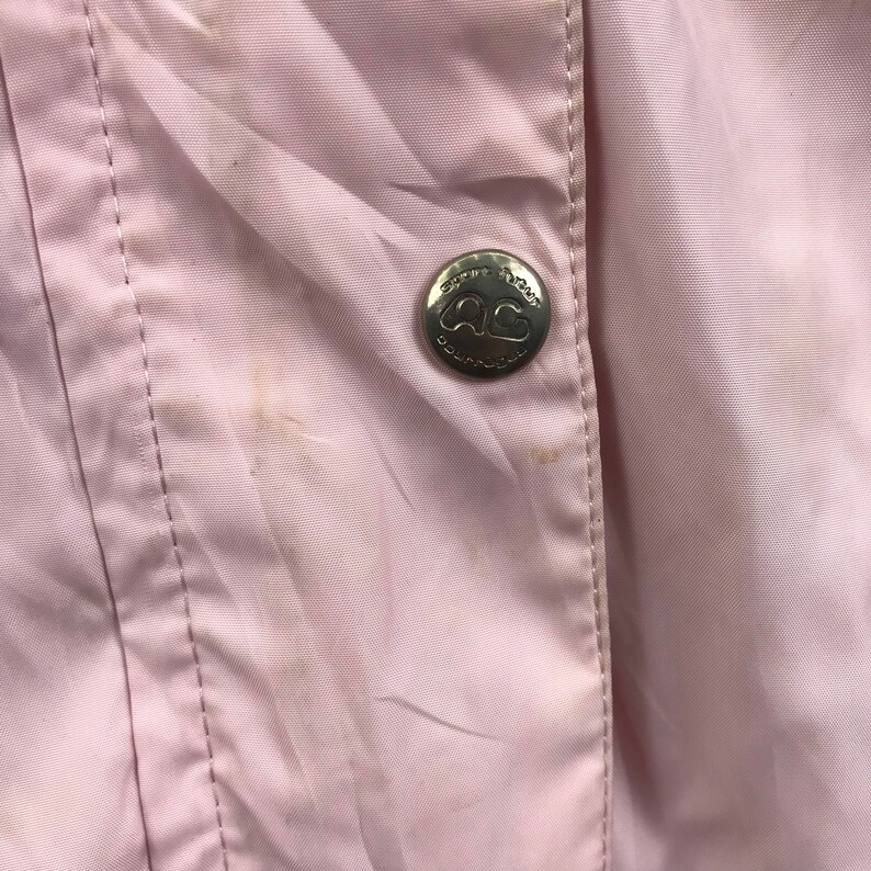 May include: Close-up of a pale pink jacket with a silver button. The button has the text "Sportswear" and the letters "AG" in a heart shape. The jacket has a vertical seam and some stains.