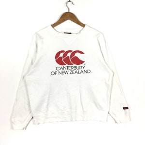 May include: A white long-sleeve crewneck sweatshirt featuring a red Canterbury logo and the black text CANTERBURY OF NEW ZEALAND printed on the chest. The garment is displayed on a wooden hanger and includes a small brand tag on the left sleeve.