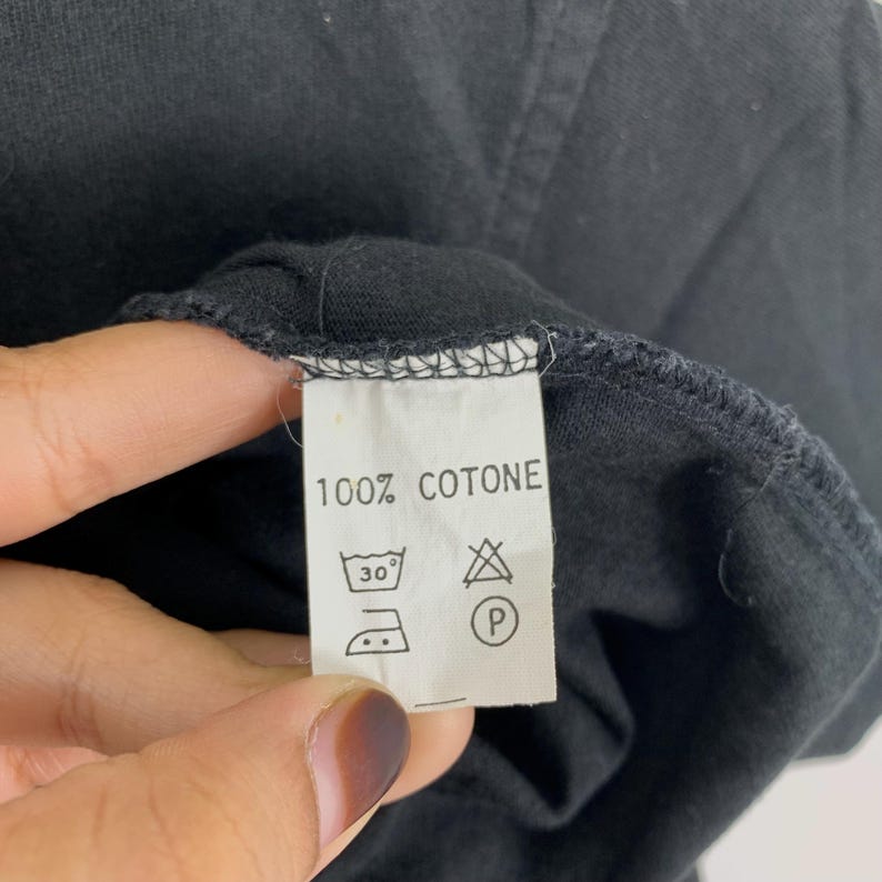 May include: Close-up of a black garment tag with the text "100% COTONE". The tag also displays washing instructions, including a 30-degree Celsius wash symbol and a dry cleaning symbol.