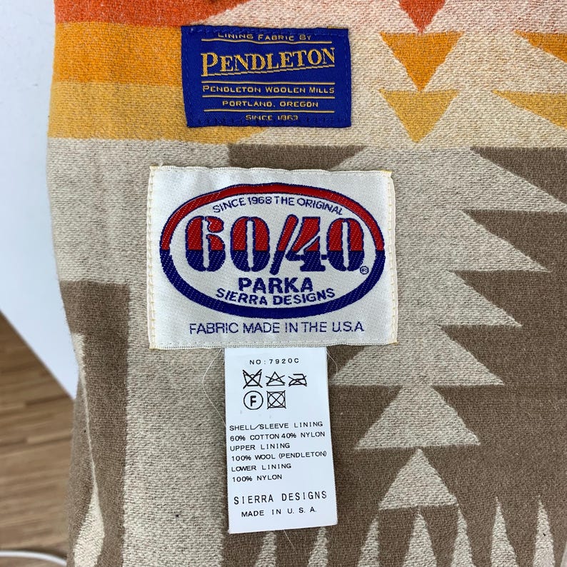 Puede incluir: Primer plano de una etiqueta de tela Pendleton y una etiqueta 60/40 Parka Sierra Designs. La etiqueta Pendleton es azul marino con texto blanco. La etiqueta 60/40 es blanca con un dise&ntilde;o ovalado rojo y azul. La tela tiene un patr&oacute;n geom&eacute;trico.