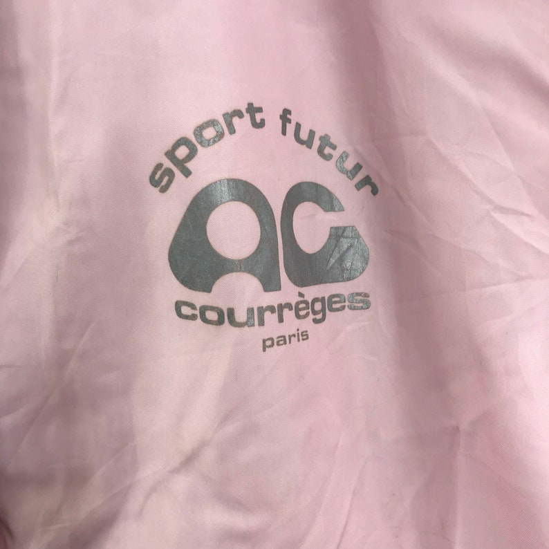 May include: A close-up of a pale pink fabric with the words "sport futur" and "courr&egrave;ges paris" printed in silver. The logo features the letters "AC" in a stylized design.