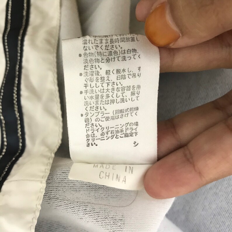May include: Close-up of a white clothing tag with Japanese text and the words "MADE IN CHINA" printed in black. The tag is attached to a white garment with a black and white stripe.