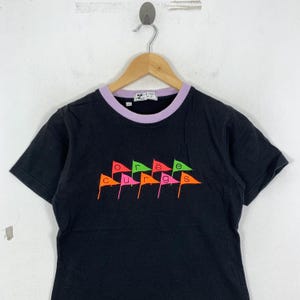 May include: Black t-shirt with a purple collar and the word "courage" spelled out in colorful flag graphics. The flags are in shades of orange, pink, green, and yellow. The shirt is hanging on a wooden hanger.