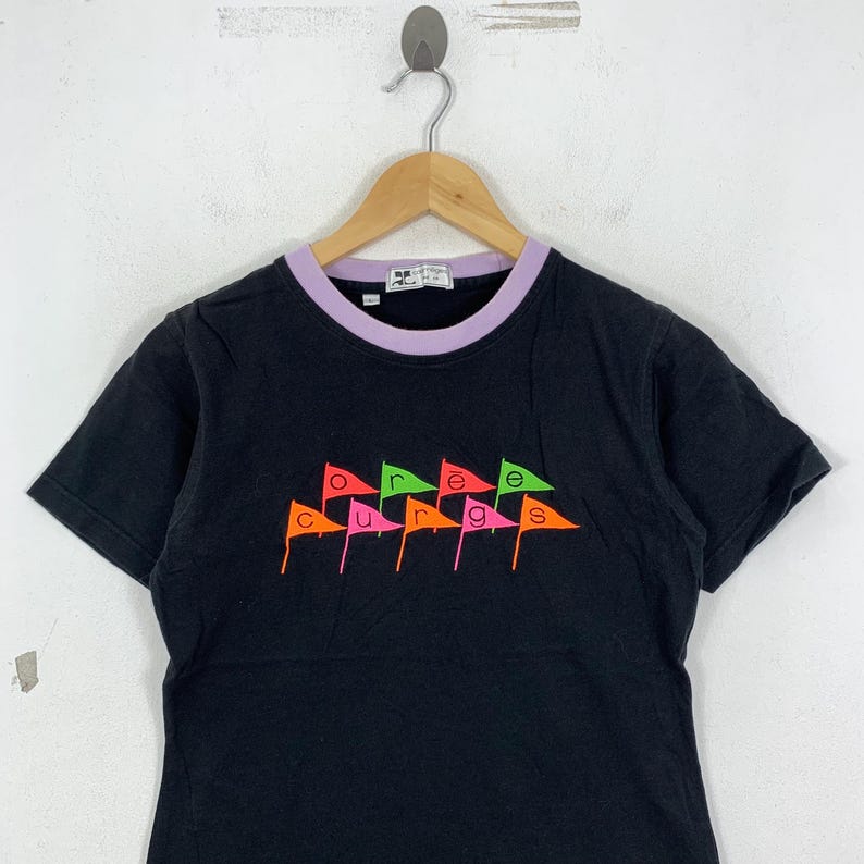 May include: Black t-shirt with a purple collar and the word "courage" spelled out in colorful flag graphics. The flags are in shades of orange, pink, green, and yellow. The shirt is hanging on a wooden hanger.