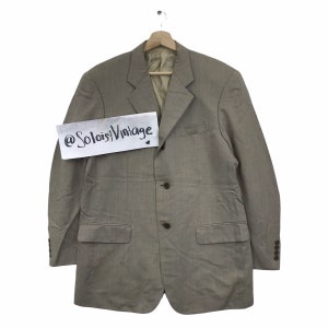 Könnte beinhalten: Beigefarbener Blazer mit Dreiknopfverschluss, zwei Vordertaschen und einem fallenden Revers. Der Blazer hängt an einem Holzkleiderbügel. Der Text "@SoloistVintage" befindet sich auf einem weißen Aufkleber.