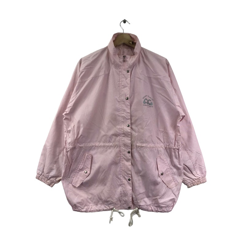 May include: A pale pink jacket with a stand-up collar, snap fastenings, and two front pockets. The jacket features a drawstring at the waist and a small logo on the upper left chest. It is hanging on a wooden hanger.