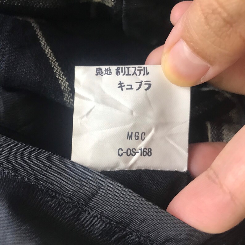 May include: A white tag with Japanese text and the letters "MGC" and "C-OS-168" printed in black. The tag is held up against a dark fabric with a subtle pattern.