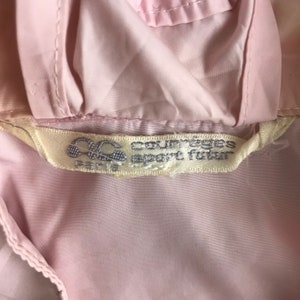 May include: A close-up shot of a pale pink garment, featuring a label with the text "Courreges sport futur" and the logo "OC PARIS". The label is off-white. The garment's fabric appears smooth and possibly synthetic.