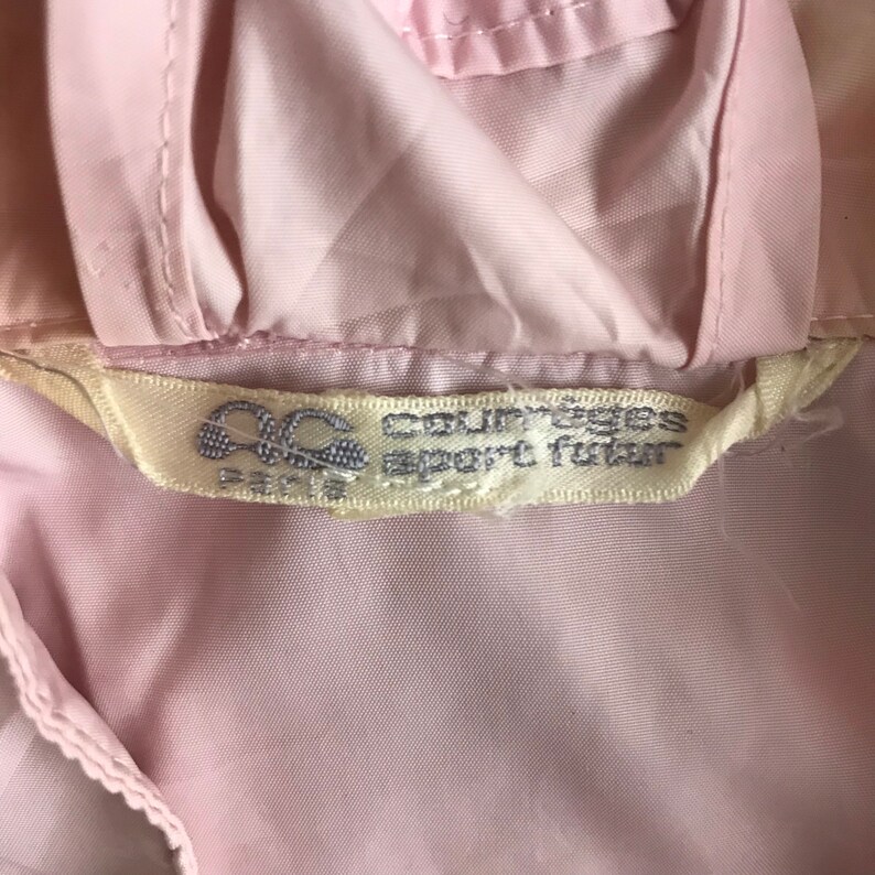 May include: A close-up shot of a pale pink garment, featuring a label with the text "Courreges sport futur" and the logo "OC PARIS". The label is off-white. The garment's fabric appears smooth and possibly synthetic.