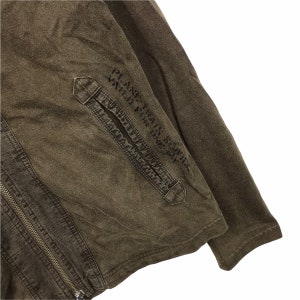 May include: Close-up of a brown jacket with a zipper closure and a pocket. The jacket has the text "PLANE TRAIN SERVICE VALID FOR ONE WAY" printed on the sleeve. The fabric appears to be a worn, textured material.