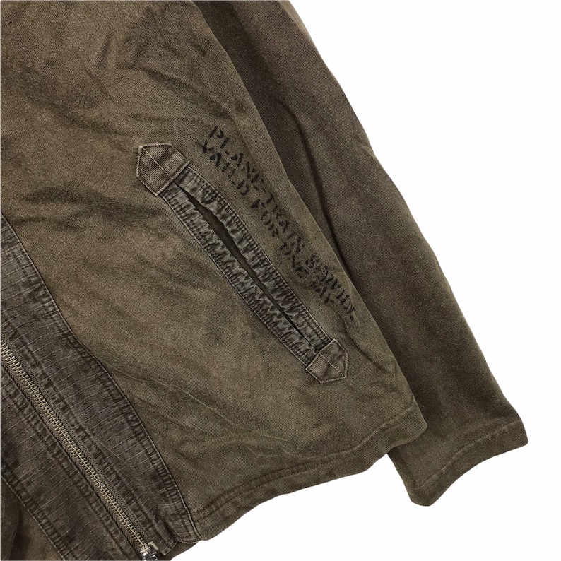 May include: Close-up of a brown jacket with a zipper closure and a pocket. The jacket has the text "PLANE TRAIN SERVICE VALID FOR ONE WAY" printed on the sleeve. The fabric appears to be a worn, textured material.