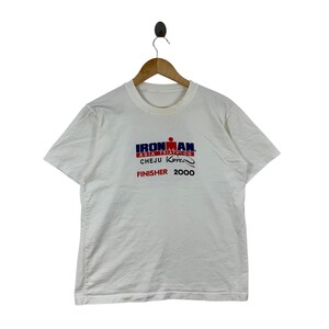 K&ouml;nnte beinhalten: Ein wei&szlig;es T-Shirt mit kurzen &Auml;rmeln und einem grafischen Aufdruck mit der Aufschrift IRONMAN ASIA TRIATHLON CHEJU KOREA FINISHER 2000. Der Text ist in blauen, roten und schwarzen Buchstaben auf der Brustmitte zu sehen, h&auml;ngend an einem Holzkleiderb&uuml;gel.
