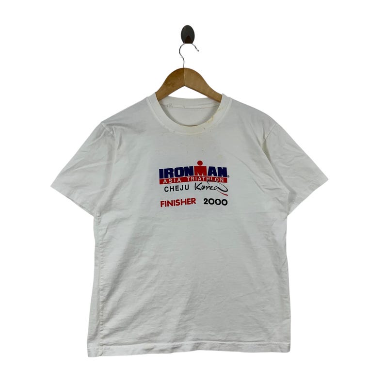 K&ouml;nnte beinhalten: Ein wei&szlig;es T-Shirt mit kurzen &Auml;rmeln und einem grafischen Aufdruck mit der Aufschrift IRONMAN ASIA TRIATHLON CHEJU KOREA FINISHER 2000. Der Text ist in blauen, roten und schwarzen Buchstaben auf der Brustmitte zu sehen, h&auml;ngend an einem Holzkleiderb&uuml;gel.