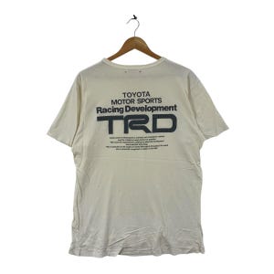 May include: Cream-colored t-shirt with the text "TOYOTA MOTOR SPORTS Racing Development" and the letters "TRD" in dark gray. The shirt is hanging on a wooden hanger.