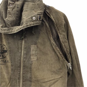 May include: Olive green military-style jacket with a hood. The jacket features a zipper closure, multiple pockets, and a patch with text and a graphic. The hood has text printed on it. The jacket is made of a durable, textured fabric.