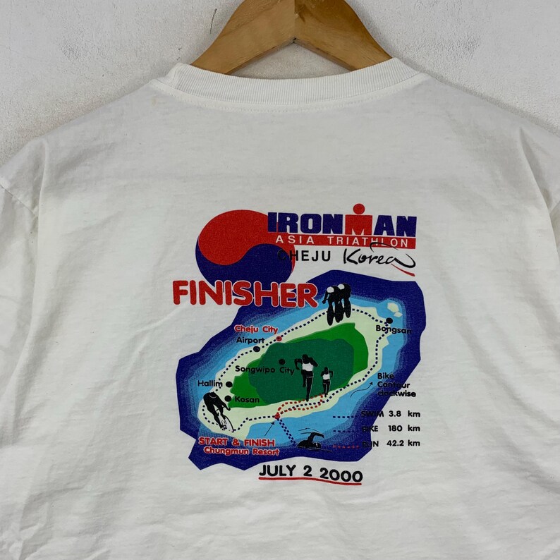 K&ouml;nnte beinhalten: Ein wei&szlig;es T-Shirt mit einer Grafik auf der R&uuml;ckseite f&uuml;r den Ironman Asia Triathlon Cheju Korea. Das Design zeigt eine Karte der Insel, Rennstatistiken, den Text FINISHER und das Datum JULY 2 2000 sowie Illustrationen von Athleten.