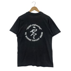 May include: Black t-shirt with a white graphic on the back. The graphic includes the text "ZERO engineering" and "WE SUPPORT FAR-EAST BIKERS" in a circular design. The shirt is hanging on a wooden hanger.