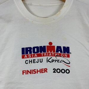 K&ouml;nnte beinhalten: Ein wei&szlig;es kurz&auml;rmeliges T-Shirt mit einem Grafikdruck, der IRONMAN ASIA TRIATHLON CHEJU KOREA FINISHER 2000 liest. Das Design enth&auml;lt ein rotes und blaues Logo mit einer stilisierten Figur. Der Kragenbereich weist sichtbare hellbraune Flecken auf.