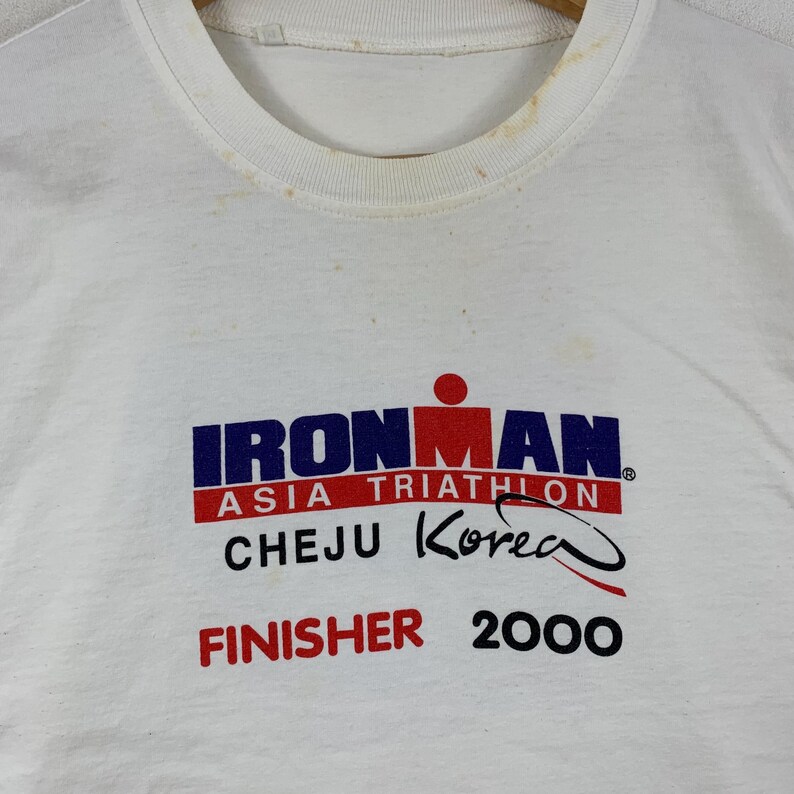 K&ouml;nnte beinhalten: Ein wei&szlig;es kurz&auml;rmeliges T-Shirt mit einem Grafikdruck, der IRONMAN ASIA TRIATHLON CHEJU KOREA FINISHER 2000 liest. Das Design enth&auml;lt ein rotes und blaues Logo mit einer stilisierten Figur. Der Kragenbereich weist sichtbare hellbraune Flecken auf.