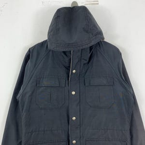 Puede incluir: Chaqueta negra con capucha, dos bolsillos en el pecho y cierres de bot&oacute;n a presi&oacute;n plateados. La chaqueta tiene una capucha con cord&oacute;n y un forro de color claro. La chaqueta est&aacute; hecha de un material liso, posiblemente resistente al agua.