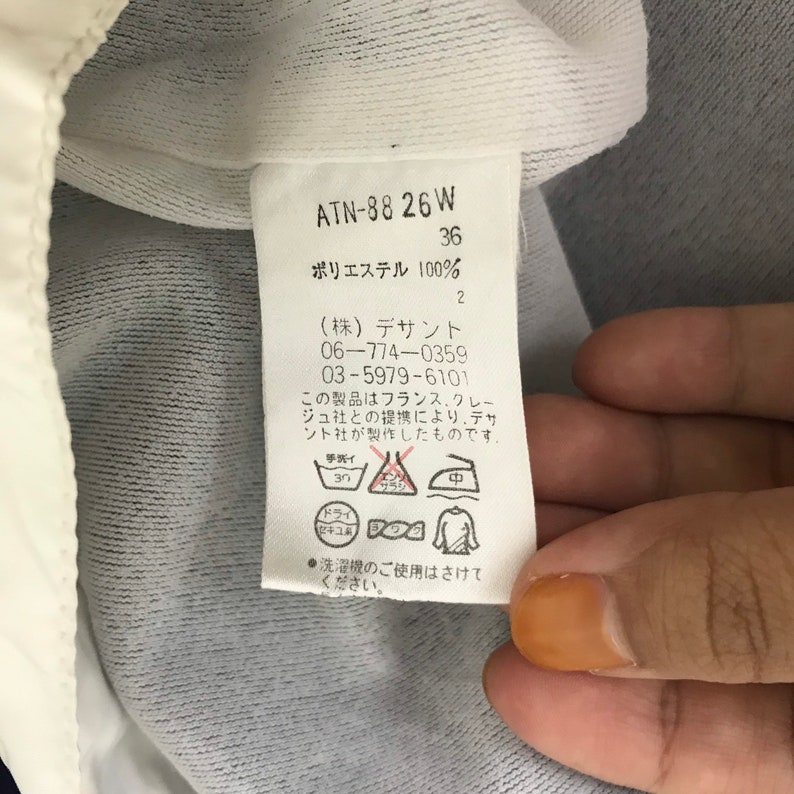 May include: A white tag with black text and symbols. The tag includes the text "ATN-88 26 W", "36", and "ポリエステル 100%", along with washing instructions and the company name. The tag is attached to a white and blue garment.