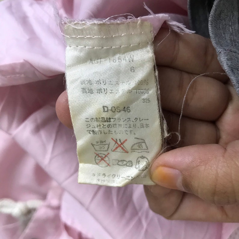 May include: A close-up of a clothing label with Japanese text and care symbols. The label is off-white with black text and is attached to a pink garment. The label includes the text "AGF-1854W" and "D-OS-46".