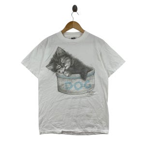 May include: A white short-sleeve t-shirt featuring a graphic illustration of a dark gray kitten sleeping inside a light blue bowl. The bowl has the word DOG printed in large blue letters on the front. The shirt is displayed on a wooden hanger.