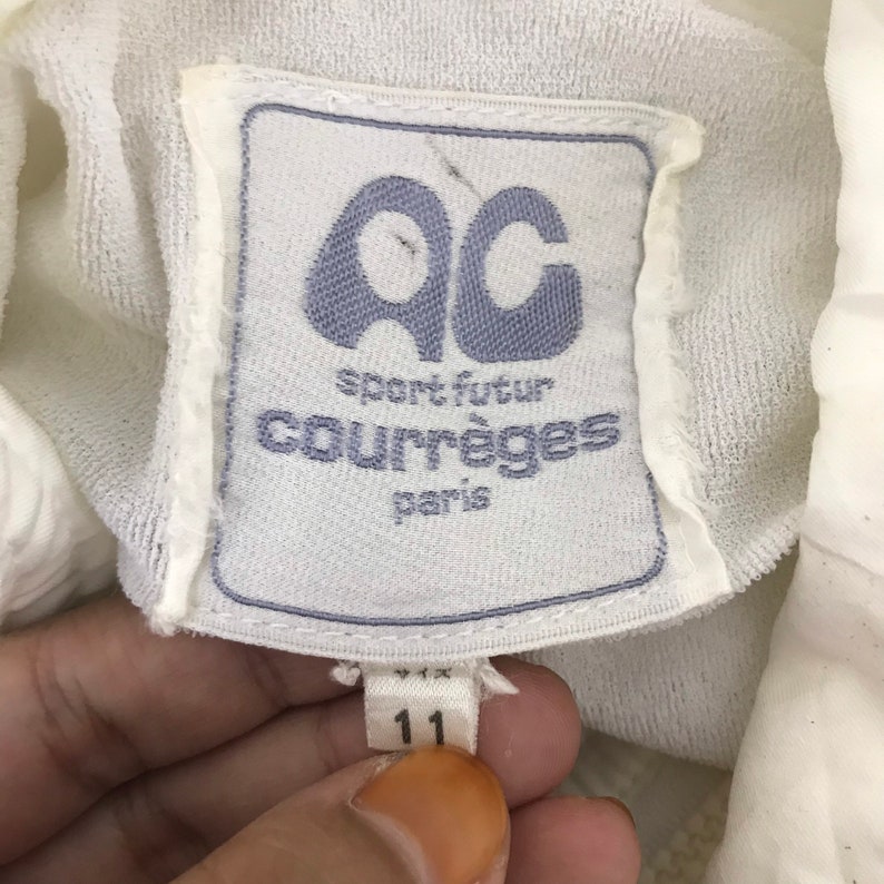 May include: A close-up of a white label with the text "AC sport futur courr&egrave;ges paris" in blue. The label also has the number "11" printed on it. The label is attached to a white fabric.