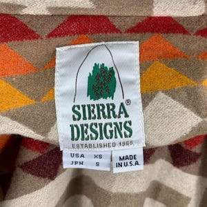 Puede incluir: Primer plano de una etiqueta Sierra Designs sobre una tela estampada. La etiqueta es blanca con texto verde y marr&oacute;n y un logotipo de monta&ntilde;a. La tela tiene un dise&ntilde;o geom&eacute;trico en rojo, naranja, amarillo y beige. La etiqueta dice "ESTABLISHED 1965" y "MADE IN U.S.A."