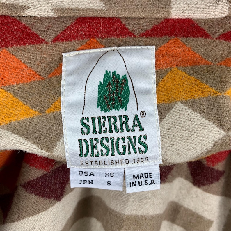 Puede incluir: Primer plano de una etiqueta Sierra Designs sobre una tela estampada. La etiqueta es blanca con texto verde y marr&oacute;n y un logotipo de monta&ntilde;a. La tela tiene un dise&ntilde;o geom&eacute;trico en rojo, naranja, amarillo y beige. La etiqueta dice "ESTABLISHED 1965" y "MADE IN U.S.A."