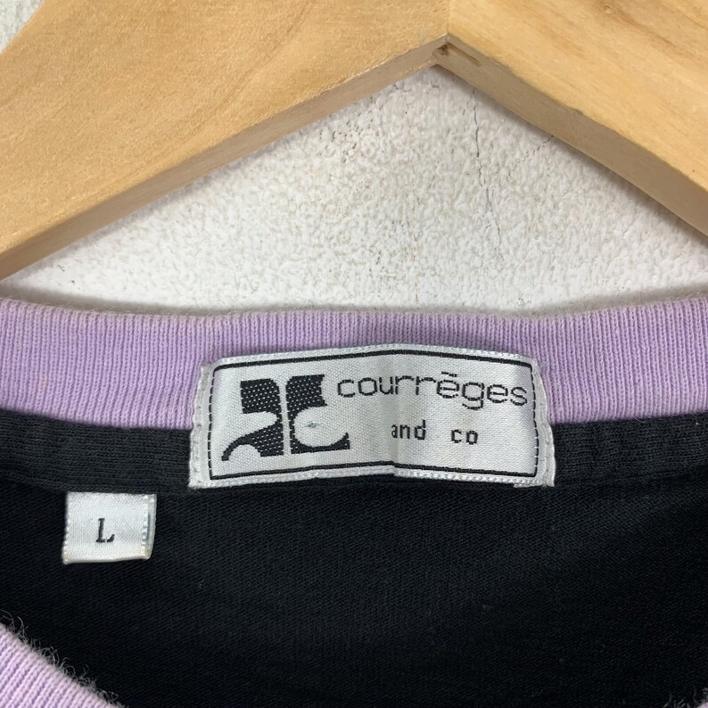 May include: Close-up of a black shirt with a lavender collar. The shirt features a white label with the text "courr&egrave;ges and co" and a size "L" tag. The shirt is hanging on a wooden hanger.