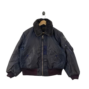 May include: A dark navy blue bomber jacket featuring a dark gray faux fur collar, a central metal zipper, and ribbed cuffs and hem. The jacket includes a small circular patch on the left sleeve, a utility pocket, and two front pockets with blue snap buttons.