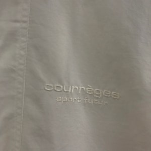 May include: A close-up of a white garment featuring the embroidered text "courr&egrave;ges sport futur" in white thread. The fabric has a slight sheen and a vertical seam detail.