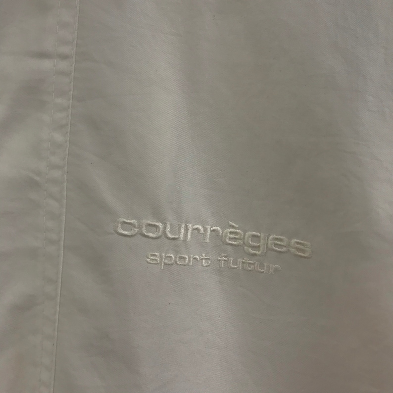 May include: A close-up of a white garment featuring the embroidered text "courr&egrave;ges sport futur" in white thread. The fabric has a slight sheen and a vertical seam detail.