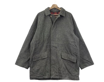 Può includere: Un cappotto grigio in lana dal design classico, con collo, due tasche frontali e chiusura con bottoni. Il cappotto è appeso a una gruccia di legno. La fodera interna presenta un motivo a quadri rossi e neri.