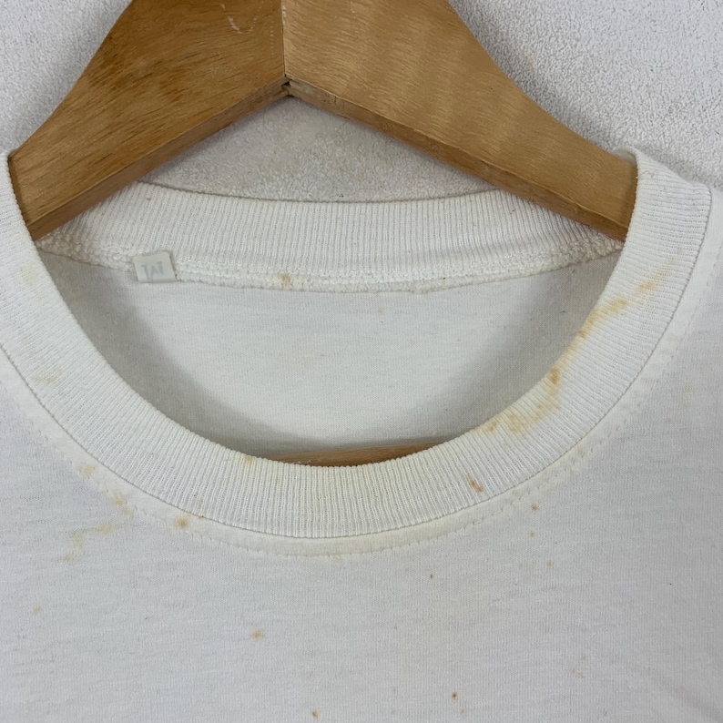 K&ouml;nnte beinhalten: Ein wei&szlig;es T-Shirt mit Rundhalsausschnitt, geripptem Kragen und einem kleinen wei&szlig;en Innenetikett mit dem Buchstaben M. Der Stoff weist sichtbare hellbraune Verf&auml;rbungen und Flecken im Bereich des Halsausschnitts auf und h&auml;ngt an einem Holzkleiderb&uuml;gel.