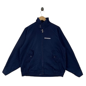 May include: A navy blue zip-up jacket featuring a high collar with two buttons, long sleeves, and two front pockets with buttoned flaps. The word SUZUKI and the Suzuki logo are embroidered in white on the upper left chest area of the garment.