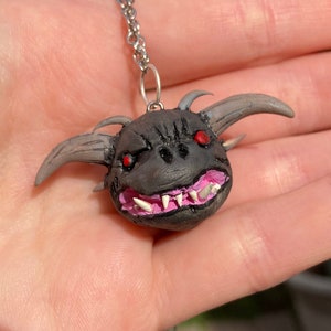 Op de afbeelding: Een ketting met een hanger van zwart en grijs klei met een klein, gedetailleerd monster met rode ogen en roze tanden. Het monster heeft hoorns en een stekelig hoofd.