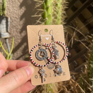 May include: A pair of handmade hoop earrings with red and white beads. The earrings feature charms of a clock, a teapot, a key, and a figure. The card reads "Handmade".