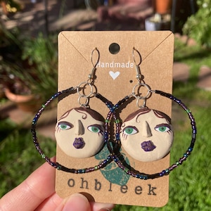 May include: Handmade hoop earrings featuring a circular face design with green eyes and purple lips. The earrings are attached to beaded hoops with a mix of purple, green, and blue beads. The earrings hang from silver-colored hooks. The card reads "ohbleek".