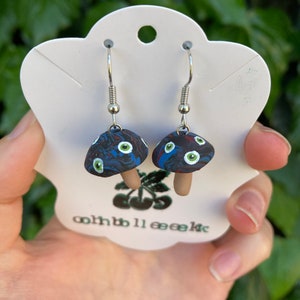 Può includere: Un paio di orecchini pendenti d'argento con ciondoli a forma di fungo blu e nero. I funghi hanno dettagli di occhi bianchi e verdi e accenti rossi.
