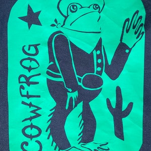 May include: A green and black graphic design of a frog wearing a cowboy hat and holding a lasso. The frog is standing on a green background with a star and the word "COWFROG" written vertically on the left side. A cactus is on the right side of the frog.