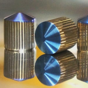 May include: Four cylindrical metal objects with a gold finish and vertical grooves. The tops are a metallic blue, and the objects are reflected on a shiny surface. The objects appear to be decorative or functional hardware.