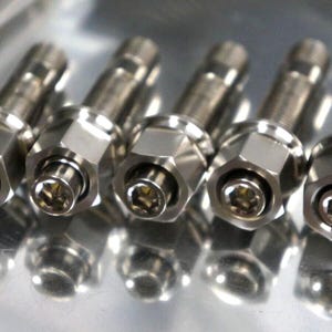 May include: Close-up of several shiny, silver metal lug bolts. Each bolt has a hexagonal head and a threaded shaft. The bolts are arranged in a row, reflecting light.