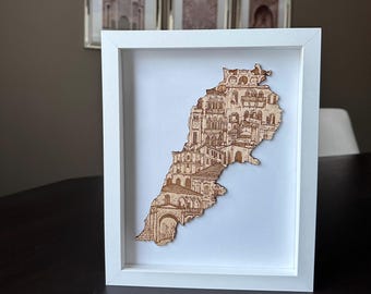 Könnte beinhalten: Eine gerahmte Holzkarte des Libanon, detailliert mit architektonischen Illustrationen. Die Karte, in hellem Braun, ist vor einem weißen Hintergrund in einem weißen Rahmen platziert. Das Kunstwerk zeigt die Wahrzeichen und Gebäude des Landes.