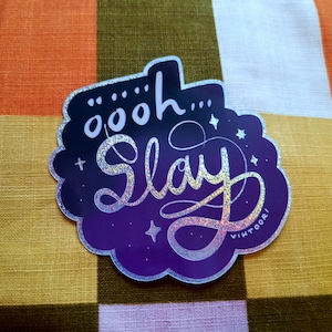 Puede incluir: Una pegatina holográfica con las palabras "öööoh... Slay" en cursiva. La pegatina tiene un fondo degradado de morado oscuro a morado claro con un borde festoneado y acentos de estrella. El nombre del artista, "vihtoor!", está en la parte inferior.